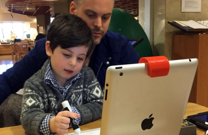 A parent and young child working on the child’s penmanship, using a tablet, marker, and something on which to write with marker. 